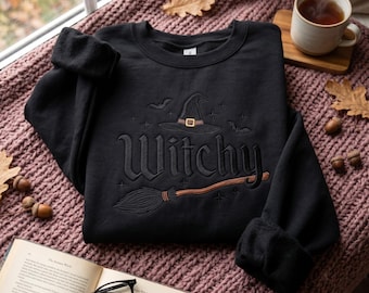 Può includere: Una felpa nera a maniche lunghe con un ricamo sul petto. Il disegno include un cappello da strega, una scopa, piccoli pipistrelli e la parola "Witchy" in un carattere stilizzato. La felpa presenta un girocollo e polsini a coste.