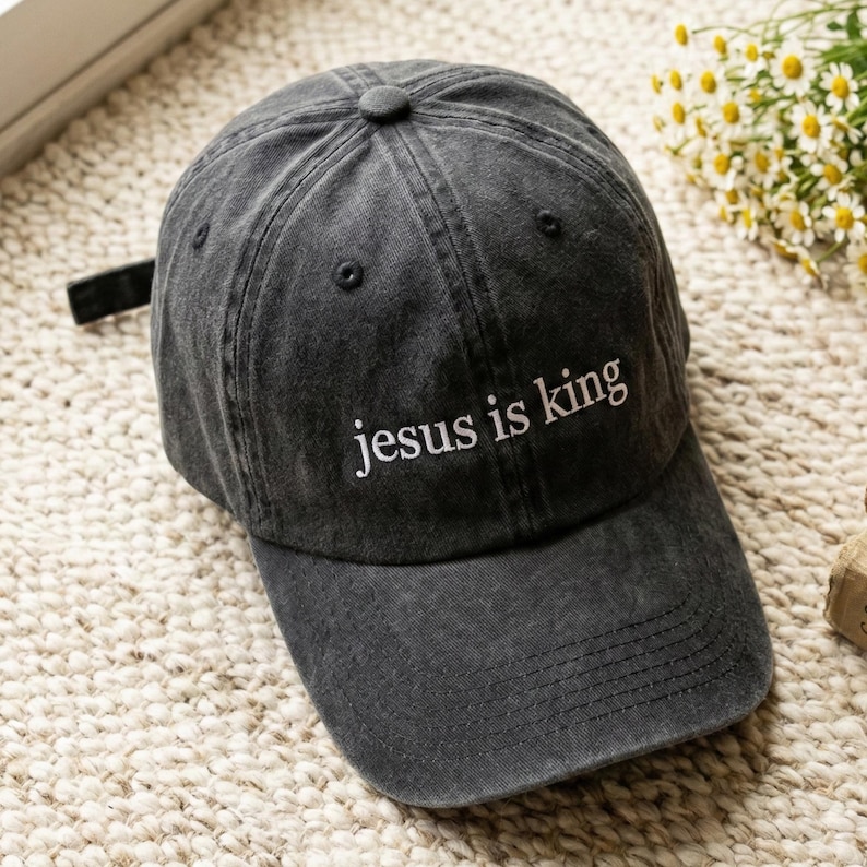 May include: A charcoal gray baseball cap with the embroidered text "jesus is king" in white. The cap has a curved brim and an adjustable strap at the back. The hat is made of a soft, worn fabric.