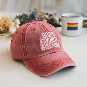 May include: A dusty rose baseball cap with the embroidered text "GOD'S FAVORITE BISEXUAL" in white. A white mug with a rainbow flag is in the background. The cap is made of a soft, worn material.