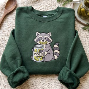 Puede incluir: Una sudadera de cuello redondo en color verde bosque oscuro con un bordado de un mapache gris y blanco que sostiene un frasco de vidrio lleno de pepinillos verdes. El mapache tiene un pepinillo en la boca y el frasco lleva la palabra PICKLES.