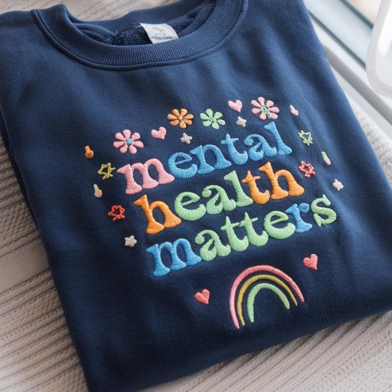 May include: Navy blue sweatshirt with the embroidered phrase "mental health matters" in colorful letters. The design includes flowers, hearts, stars, and a rainbow, creating a cheerful and supportive message.