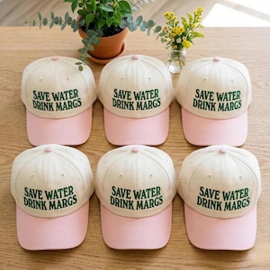 May include: Six baseball caps arranged in two rows, each featuring an off-white crown and a light pink brim. The front of every cap displays the embroidered green text "SAVE WATER DRINK MARGS" in a bold, sans-serif font with a button on top.