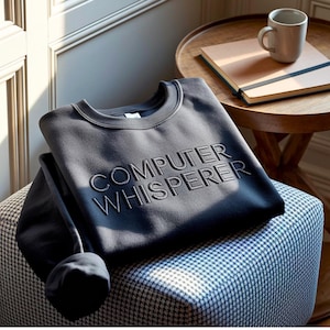 May include: A dark charcoal gray crewneck sweatshirt featuring the text "COMPUTER WHISPERER" embroidered in a matching thread color across the chest. The garment has long sleeves and a ribbed neckline, showcasing a clean and simple design.
