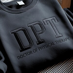 May include: Dark gray sweatshirt with the embroidered letters "DPT" above the words "DOCTOR OF PHYSICAL THERAPY". The sweatshirt is made of a soft material and has a crew neck.