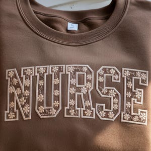 May include: A brown crewneck sweatshirt with the word "NURSE" embroidered across the front in a floral design. The letters are outlined in white and filled with small, cream-colored flowers. The sweatshirt has a ribbed neckline and cuffs.