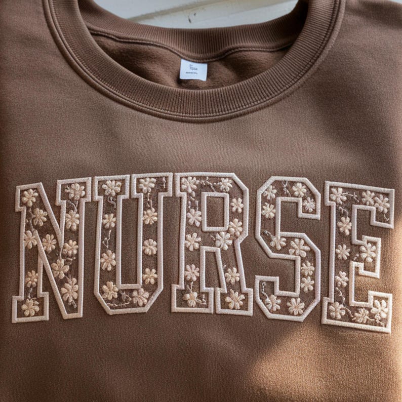 May include: A brown crewneck sweatshirt with the word "NURSE" embroidered across the front in a floral design. The letters are outlined in white and filled with small, cream-colored flowers. The sweatshirt has a ribbed neckline and cuffs.