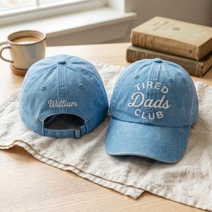 May include: Two light blue baseball caps. One cap has the text "Tired Dads Club" embroidered in white. The other cap has the name "William" embroidered in white. Both caps have an adjustable strap and a curved brim.