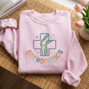 Könnte beinhalten: Ein hellrosa Sweatshirt mit dem Wort "ORTHOPEDICS" in bunten Buchstaben gestickt. Das Design beinhaltet ein Kreuz mit einer Knie-Illustration. Die Ärmel sind gerollt. Ein Buch und Blumen sind im Hintergrund.