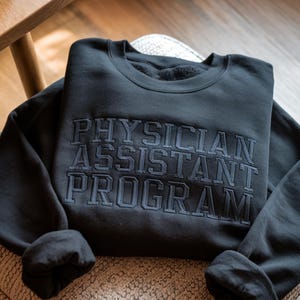 May include: A dark gray sweatshirt with the embroidered text "PHYSICIAN ASSISTANT PROGRAM" in a matching dark gray color. The sweatshirt is folded and resting on a patterned surface.