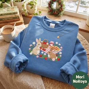 May include: A blue crewneck sweatshirt featuring an embroidered picnic scene with two brown rabbits, a basket of strawberries, a red-checkered blanket, a pie, and a jar of jam, surrounded by small white and pink flowers.