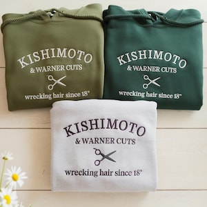 May include: Three hooded sweatshirts in olive green, dark green, and light gray. Each features embroidered text reading KISHIMOTO & WARNER CUTS and wrecking hair since 18, accompanied by a pair of scissors graphic centered on the chest area.