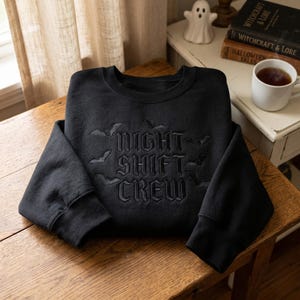 May include: A black crewneck sweatshirt featuring black embroidery that reads "NIGHT SHIFT CREW" in a gothic font, surrounded by small embroidered bat silhouettes. The garment has long sleeves and a ribbed neckline, cuffs, and hem.