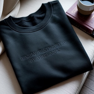 May include: Black sweatshirt with the embroidered text "EASILY DISTRACTED BY AIRPLANES." The crew neck sweatshirt is folded on a light-colored surface, with a book and a mug in the background.
