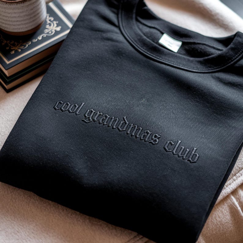 May include: Black crew neck sweatshirt with the embroidered text "cool grandmas club". The sweatshirt is folded and resting on a beige surface. A cup and a book are visible in the background.