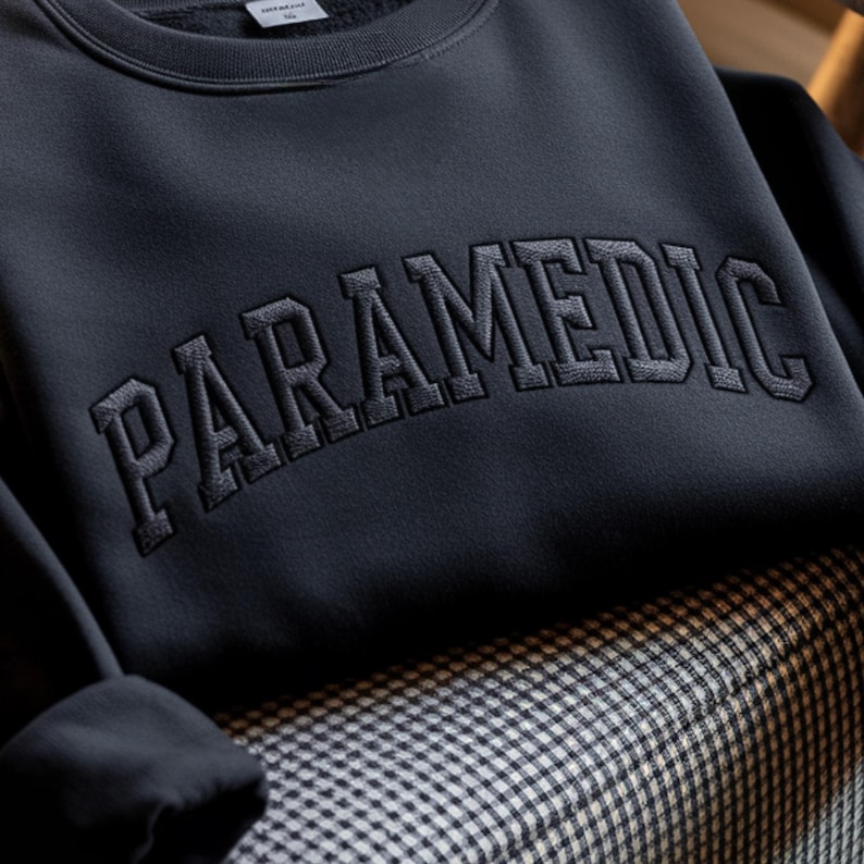 May include: A dark charcoal gray crewneck sweatshirt featuring the word PARAMEDIC embroidered in a matching dark thread across the chest in a bold, arched collegiate-style font. The garment has a ribbed neckline and long sleeves.