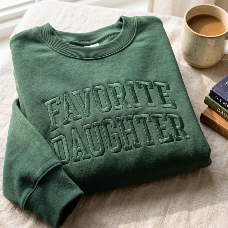 Pode incluir: Um moletom verde escuro com a frase bordada "FAVORITE DAUGHTER" em letras grossas. O moletom est&aacute; dobrado, com uma caneca de caf&eacute; e livros ao fundo. O tecido parece macio e confort&aacute;vel.