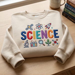 May include: A beige crewneck sweatshirt featuring colorful embroidered science-themed icons and the word SCIENCE in bold, multi-colored letters. The design includes a satellite, atom, rocket, microscope, test tubes, magnifying glass, plant, and molecule.