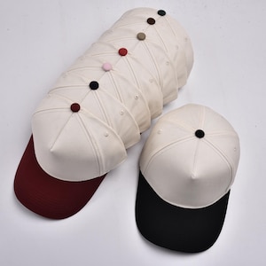 Op de afbeelding: Een verzameling baseballpetten met witte kronen en gekleurde kleppen. De petten hebben een knoop bovenop en zijn in een groep gerangschikt, met &eacute;&eacute;n pet apart. Kleuren van de klep zijn bordeauxrood en zwart.