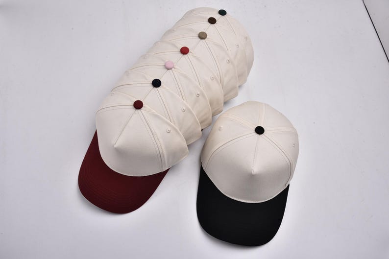 Op de afbeelding: Een verzameling baseballpetten met witte kronen en gekleurde kleppen. De petten hebben een knoop bovenop en zijn in een groep gerangschikt, met &eacute;&eacute;n pet apart. Kleuren van de klep zijn bordeauxrood en zwart.