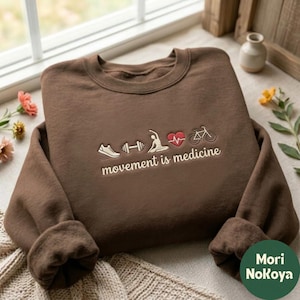May include: A brown crewneck sweatshirt featuring embroidered icons of a running shoe, a dumbbell, a person stretching, a red heart with a pulse line, and a bicycle, with the text "movement is medicine" stitched below in white.