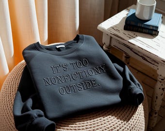 May include: A dark charcoal gray crewneck sweatshirt featuring the embroidered text IT'S TOO NONFICTIONY OUTSIDE. The garment has long sleeves with ribbed cuffs and a ribbed neckline, constructed from a solid-colored fabric with a smooth finish.