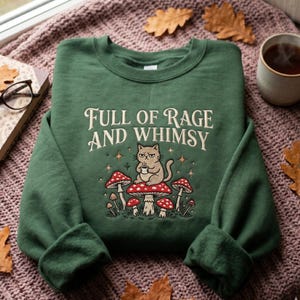 May include: A dark green crewneck sweatshirt featuring the text "FULL OF RAGE AND WHIMSY" in cream lettering. Below the text is a graphic of a grumpy cat holding a small cup, sitting atop a large red mushroom surrounded by smaller mushrooms.