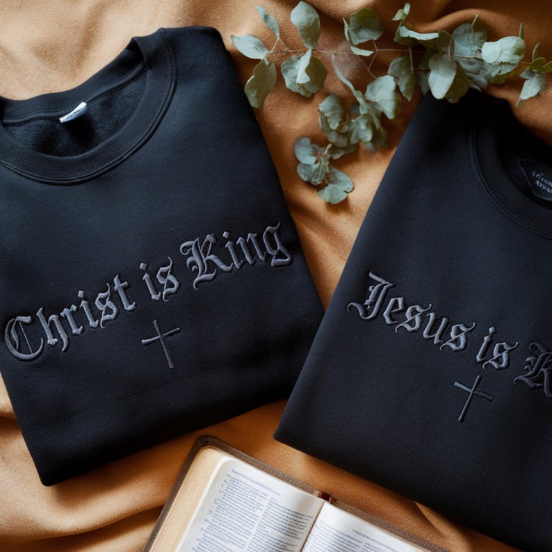 May include: Two black crewneck sweatshirts featuring embroidered text and a cross symbol. The left sweatshirt displays the text Christ is King, and the right sweatshirt displays the text Jesus is King, both written in a gothic-style font.