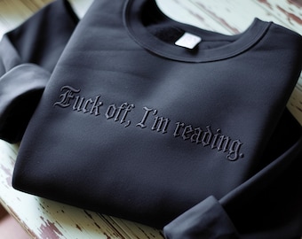 May include: Black sweatshirt with the phrase "Fuck off, I'm reading" in gothic lettering. The sweatshirt is folded on a light-colored wooden surface. The sleeves are folded up.