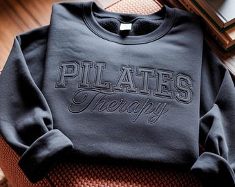 May include: A dark charcoal gray crewneck sweatshirt featuring the embroidered text PILATES Therapy on the front. The word PILATES is in a bold, block-style font, while the word Therapy is in a cursive script font, both in a matching dark thread.