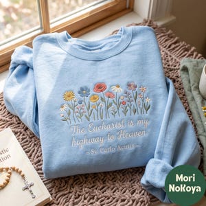 Könnte beinhalten: Ein hellblaues Sweatshirt mit Rundhalsausschnitt und einer Stickerei aus bunten Wildblumen in Gelb-, Blau-, Rosa- und Weißtönen. Unter den Blumen steht in weißer Schrift: "The Eucharist is my highway to Heaven - St. Carlo Acutis -."