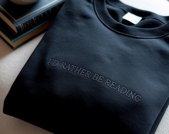 May include: A black crewneck sweatshirt featuring the embroidered text I'D RATHER BE READING in a matching black thread across the chest. The garment has a ribbed collar and a clean, solid-colored design with a white interior neck label.