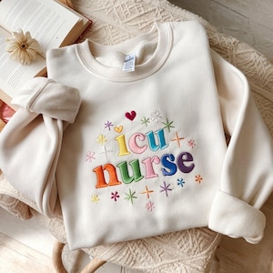 May include: Cream-coloured sweatshirt with the embroidered text "ICU Nurse" in rainbow colours. The design includes hearts, stars, and floral accents. The long-sleeved top is folded on a textured surface.