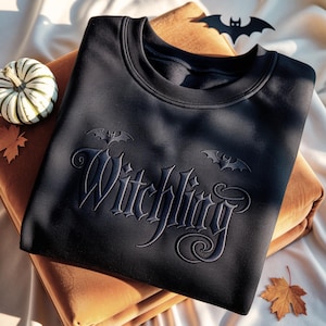 May include: A black crewneck sweatshirt featuring the word Witchling embroidered in a dark, tonal thread across the chest. The design includes two small embroidered bats positioned above the text, with a larger bat icon near the neckline.