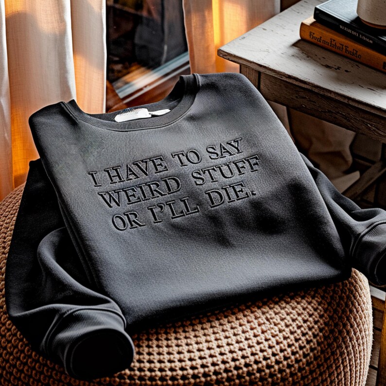 Puede incluir: Sudadera gris oscuro con la frase bordada: "I HAVE TO SAY WEIRD STUFF OR I'LL DIE". La sudadera est&aacute; doblada sobre un puf marr&oacute;n tejido. Las mangas est&aacute;n dobladas. Una etiqueta es visible en el cuello.