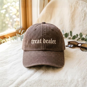 May include: A brown baseball cap with the text "treat dealer" embroidered in white. The cap is made of a soft material and has a curved brim. Sunglasses are visible in the background.