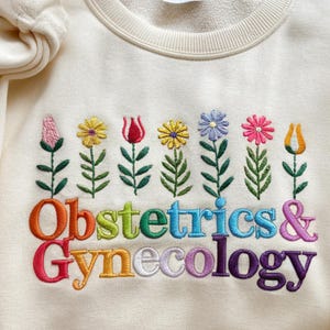 May include: Cream-colored sweatshirt embroidered with colorful flowers and the words "Obstetrics & Gynecology." The embroidery features a row of stylized flowers in various colors above the text, which is in a rainbow of hues.