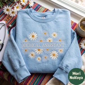 May include: A light blue crewneck sweatshirt featuring white and yellow embroidered daisy flowers surrounding the text "MEDICAL ASSISTANT" in white capital letters. The design is centred on the chest of the long-sleeved garment.