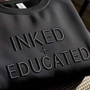 Może przedstawiać: Czarna bluza z okrągłym dekoltem, na kt&oacute;rej na piersi wyhaftowano słowa INKED + EDUCATED pasującą czarną nicią. Tekst jest wyśrodkowany i wykonany pogrubioną czcionką bezszeryfową z widoczną wypukłą fakturą haftu.