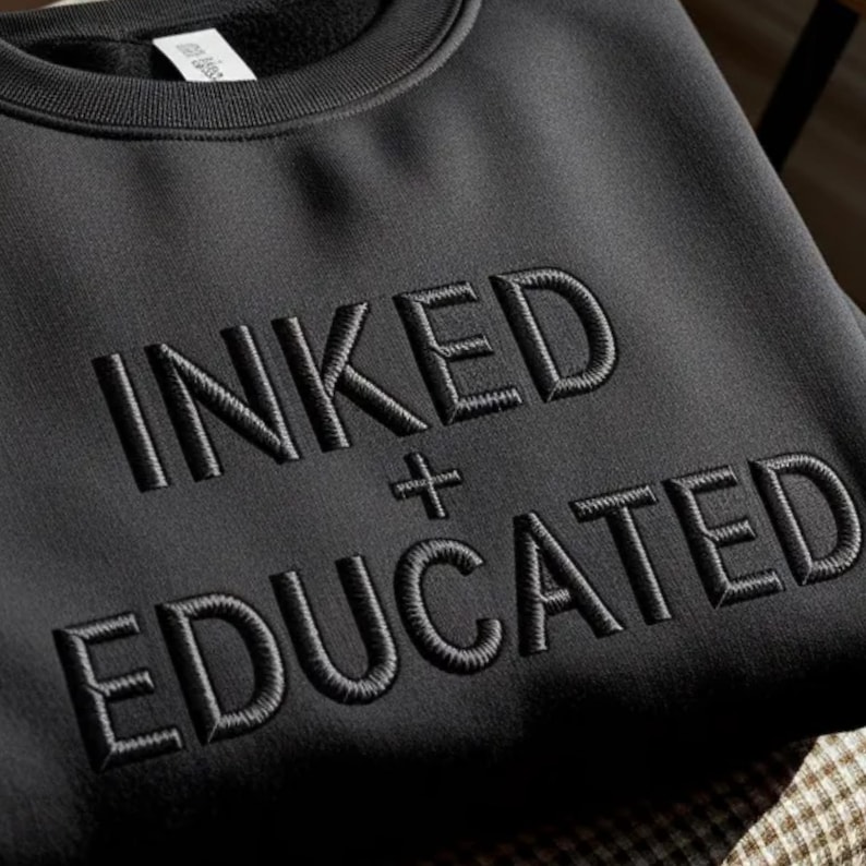 Może przedstawiać: Czarna bluza z okrągłym dekoltem, na kt&oacute;rej na piersi wyhaftowano słowa INKED + EDUCATED pasującą czarną nicią. Tekst jest wyśrodkowany i wykonany pogrubioną czcionką bezszeryfową z widoczną wypukłą fakturą haftu.