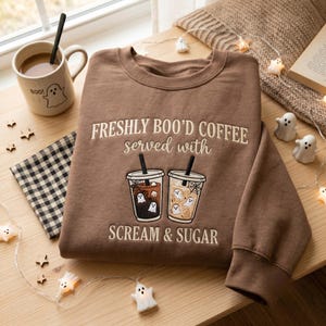 Puede incluir: Una sudadera marrón de cuello redondo con el texto bordado FRESHLY BOO'D COFFEE served with SCREAM & SUGAR, acompañada de una ilustración de dos vasos de café helado con pequeñas figuras de fantasmas blancos y pajitas negras.