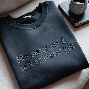 May include: A black crewneck sweatshirt featuring the embroidered text "My sister and I talk shit about you" in a dark gothic-style font. The garment has a ribbed collar and long sleeves, with the embroidery centred on the chest area.
