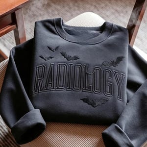 May include: A black crewneck sweatshirt featuring the word RADIOLOGY embroidered in a bold, collegiate-style font across the chest. The design is accented with several small, embroidered black bats flying around and above the lettering.