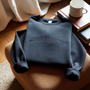 May include: A navy blue sweatshirt with the word "SONOGRAPHER" embroidered across the chest. The sweatshirt is folded on a brown woven chair. A white mug and books are in the background.