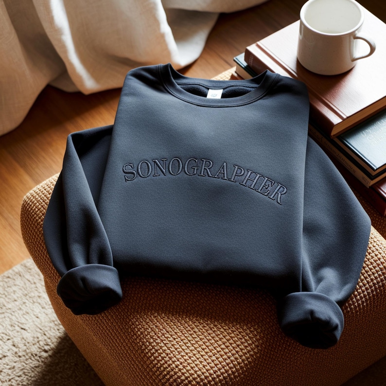 May include: A navy blue sweatshirt with the word "SONOGRAPHER" embroidered across the chest. The sweatshirt is folded on a brown woven chair. A white mug and books are in the background.