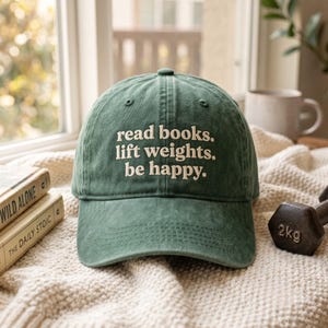 May include: A forest green baseball cap with the embroidered white text "read books. lift weights. be happy." The cap features a structured crown, a curved brim, and stitched eyelets on the top panels, presenting a classic six-panel design.