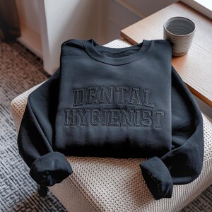 Peut inclure: Sweat-shirt noir avec l'inscription brod&eacute;e "DENTAL HYGIENIST" en police de style universitaire. Le v&ecirc;tement est pli&eacute; sur une surface &agrave; motifs. Une petite table et une tasse sont visibles en arri&egrave;re-plan.
