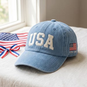 Puede incluir: Una gorra de béisbol de mezclilla azul claro con las letras USA en grandes letras blancas bordadas en la parte delantera. El lateral de la gorra muestra una pequeña bandera estadounidense bordada con el texto 1776-2026 impreso debajo.