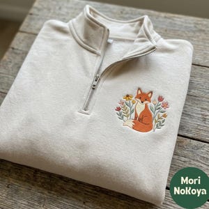 May include: A cream-colored quarter-zip sweatshirt featuring an embroidered design on the chest. The embroidery depicts an orange fox sitting amidst a cluster of colorful flowers and green leaves. The sweatshirt has a silver-toned metal zipper.