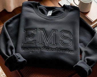 May include: A black long-sleeve crewneck sweatshirt featuring the letters EMS in a large, bold, embroidered font across the chest. Below the letters, the text Emergency Medical Services is embroidered in a smaller, cursive-style font.