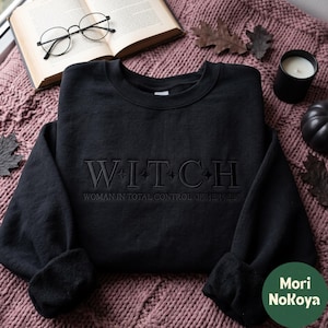 以下が含まれることがあります： 黒のクルーネックスウェットシャツ。中央に「WITCH」という刺繍があり、文字の間に小さな星のアクセントが施されています。そのすぐ下には「WOMAN IN TOTAL CONTROL OF HERSELF」というフレーズが小さめの文字で刺繍されています。